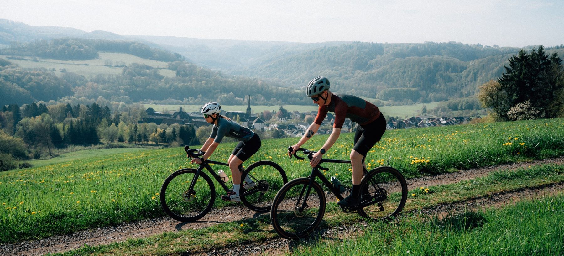 Two cyclists wearing helmets ride on a dirt trail through a lush green field with hills, forests, and a village visible in the background under a clear sky.